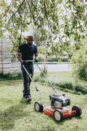 En man går bakom en gräsklippare och klipper gräset under ett träd en solig dag.