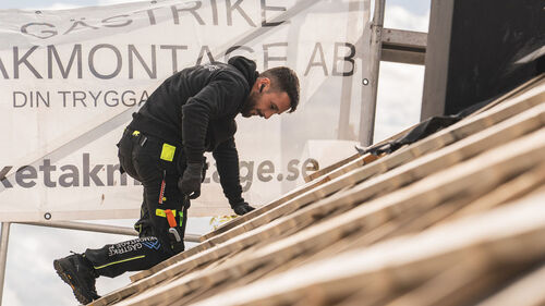 Takläggare i svart arbetskläder arbetar på ett trätak framför en stor banderoll med företagsnamnet 