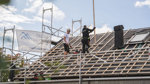 Översiktsbild av två takläggare på ett stort lutande tak, med ställning, banderoll med 