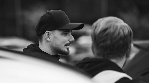 Svartvit bild av två män som pratar, med fokus på mannen i keps och arbetsjacka från Gästrike Takmontage AB.