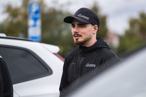 En man i svart jacka och keps med Gästrike Takmontage-logotyp står utomhus vid vita bilar.