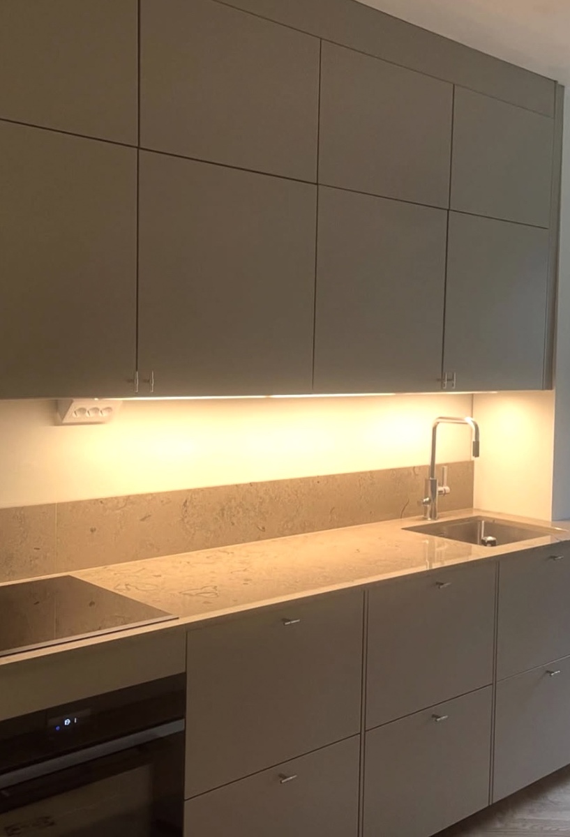 Nyrenoverat kök med moderna släta skåpsluckor, bänkskiva i sten och installerad belysning under överskåpen.