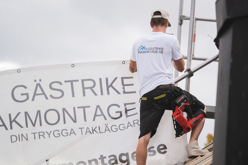 Takläggare i vit t-shirt med Gästrike Takmontage AB-logga arbetar vid ett takräcke intill företagets banderoll.