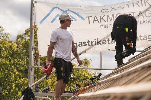 Två takläggare från Gästrike Takmontage AB arbetar på ett tak, med en stor banderoll i bakgrunden.