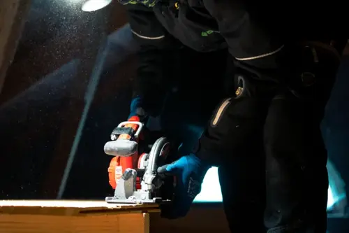 Person i arbetskläder som använder cirkelsåg för att såga trä i ett mörkt utrymme med damm i luften.