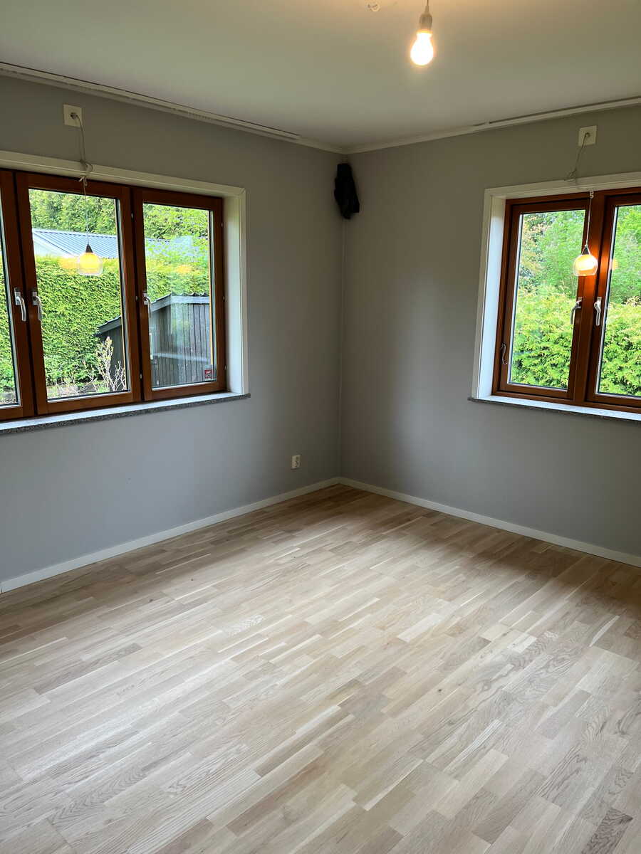 Ljust rum med nyslipat trägolv och fönster med utsikt mot trädgård.