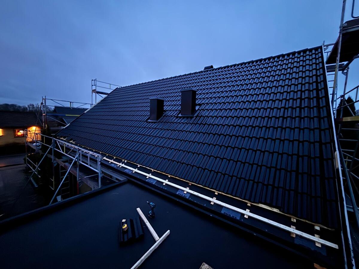 Färdiglagt tak med svarta betongtakpannor och ventilationshuvar fotograferat i skymning