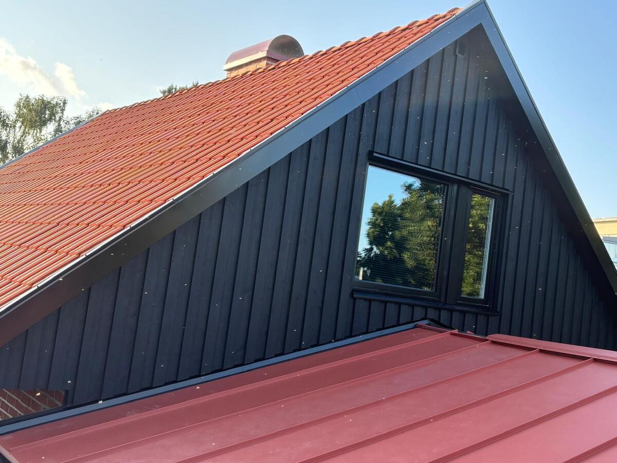 Modernt hus med röd stående falsad plåt och svart träfasad med takfönster i soligt väder