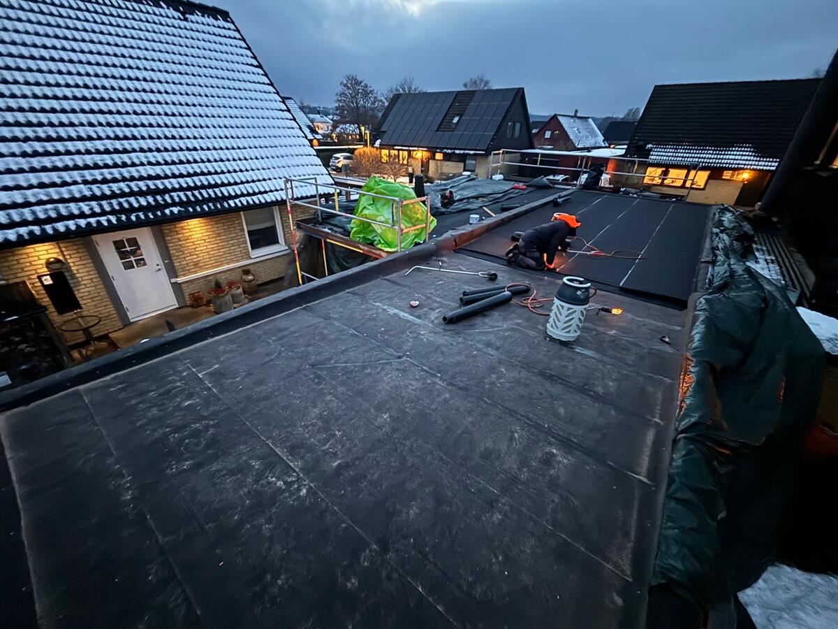 Takläggare arbetar med tätskiktsmatta på platt tak i skymning med angränsande villatak