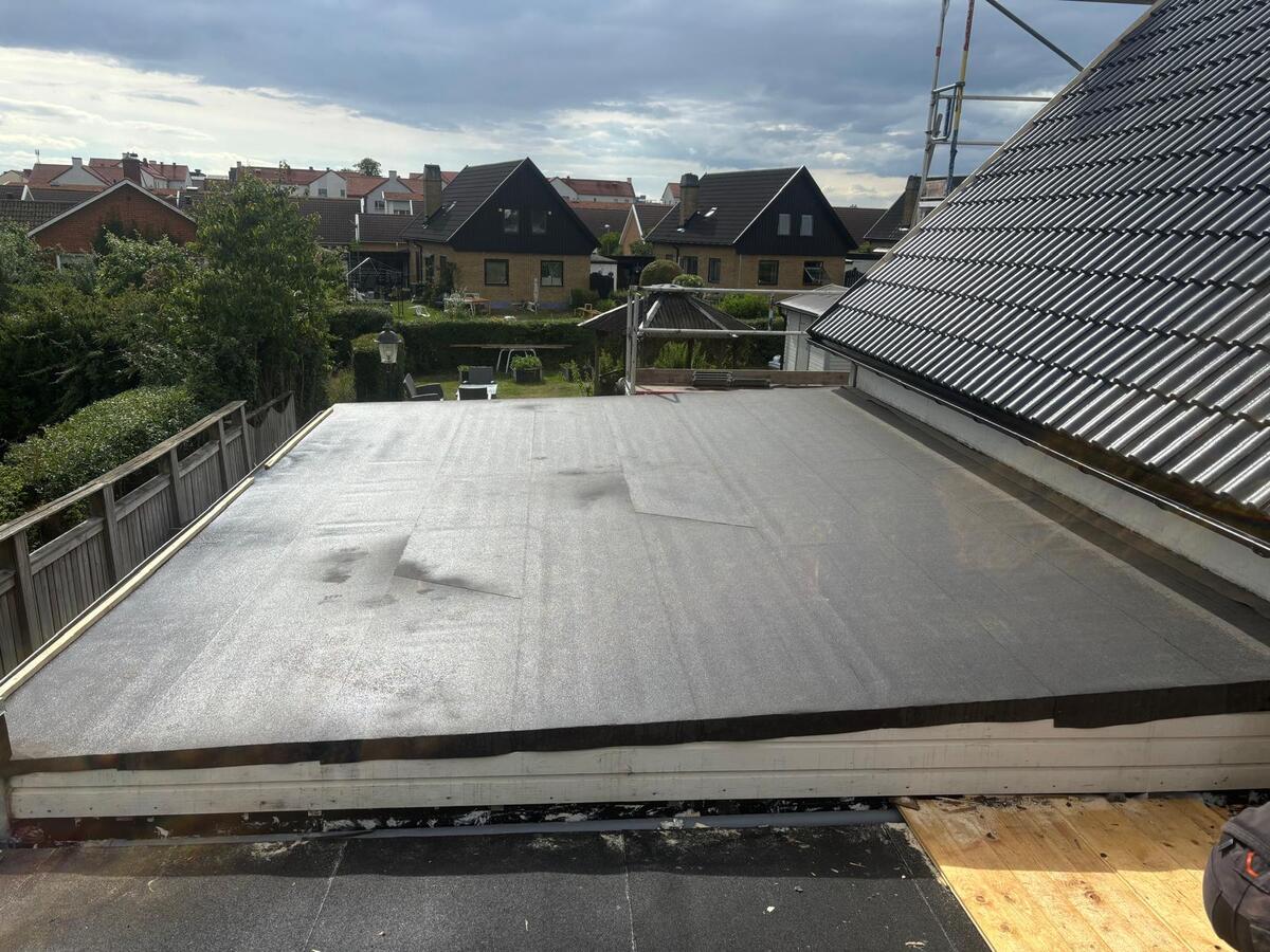 Färdiglagt platt tak med grå tätskiktsmatta på villa med svart tegeltak i skånskt villaområde