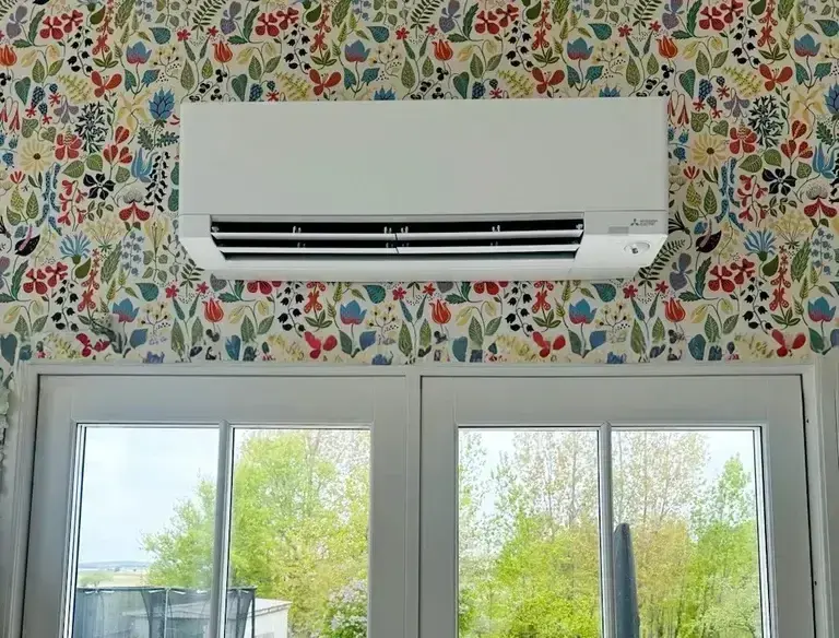 Vit luftvärmepump monterad ovanför fönster med blommig tapet