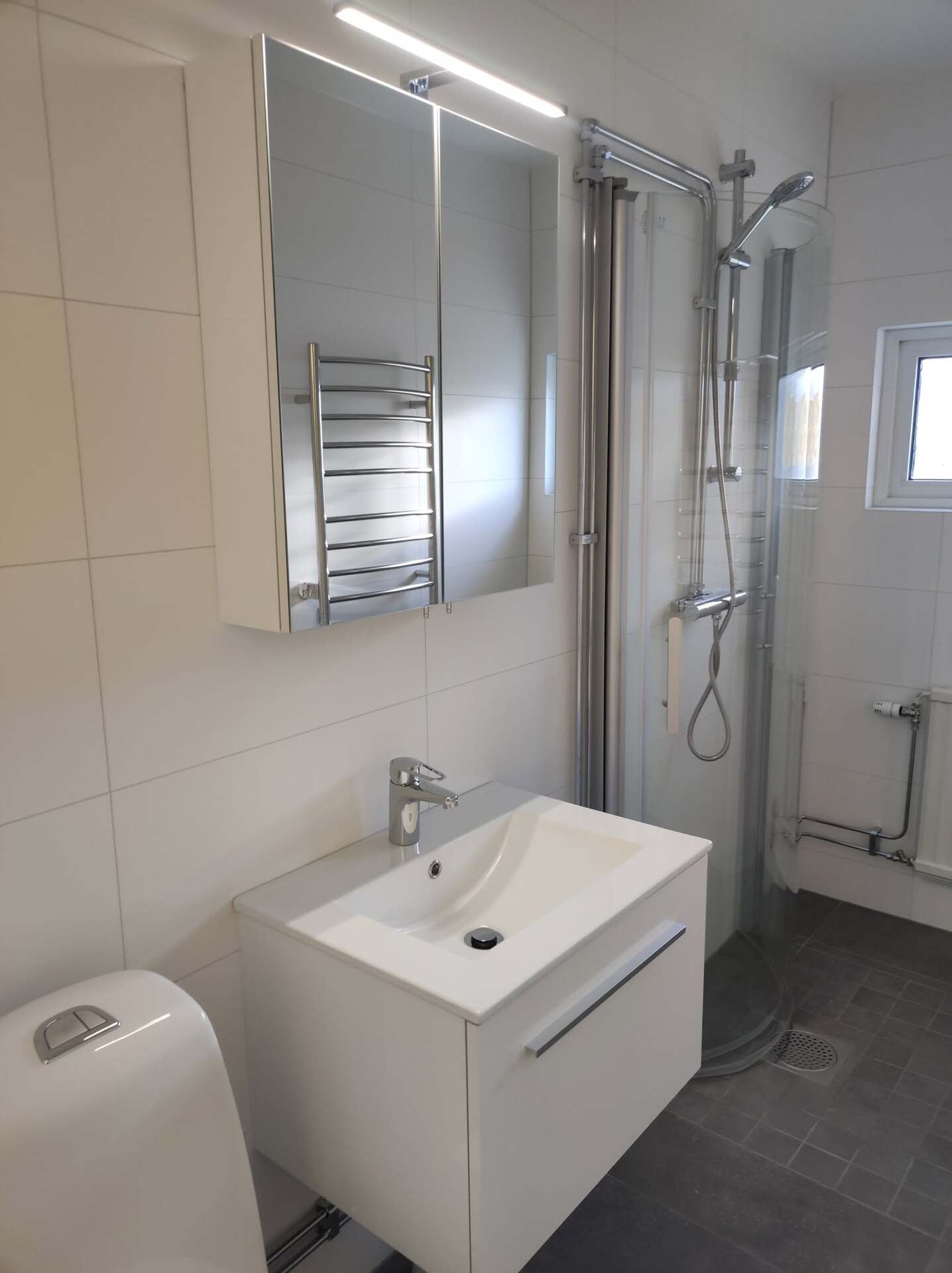 Renoverat badrum med vit tvättställ, glasad dusch och handdukstork i krom