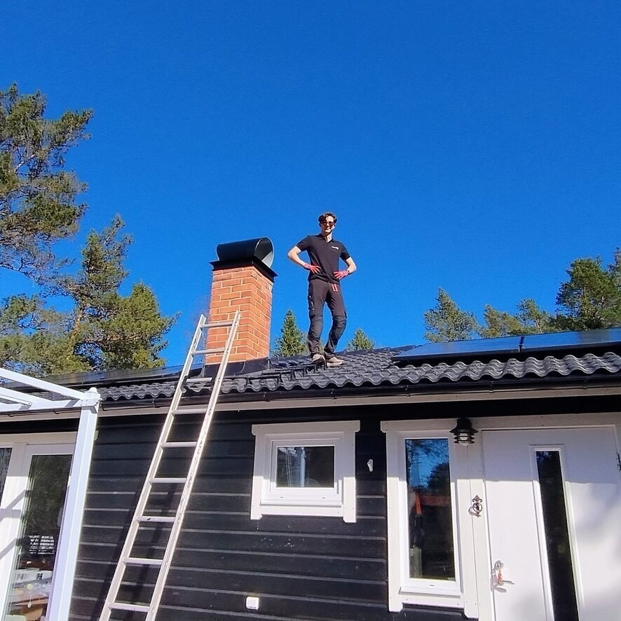Montör som står vid tegelskorsten på tak, med stege och klarblå himmel