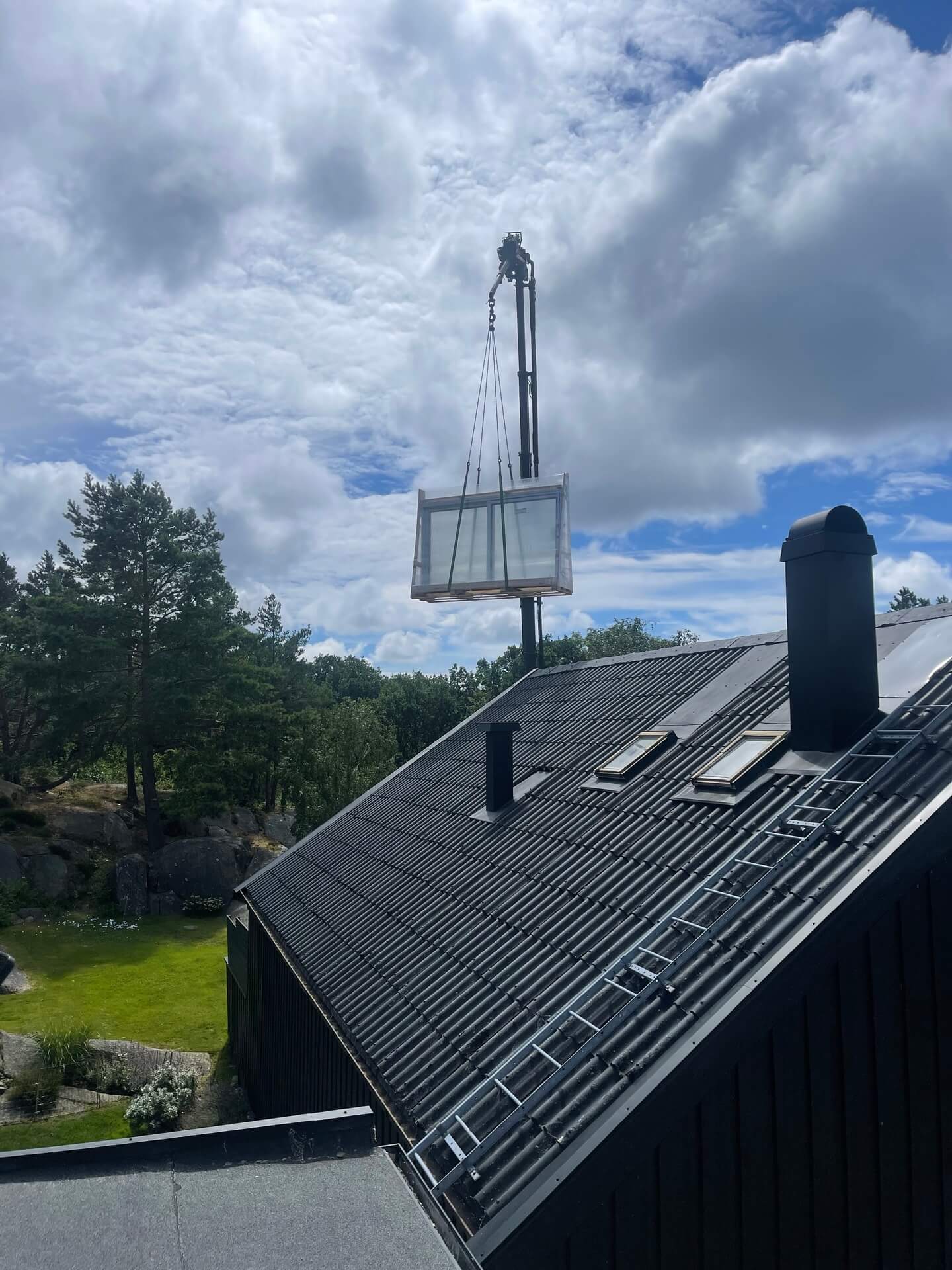Fönsterparti lyfts med kran över mörkt tegeltäckt tak vid installation