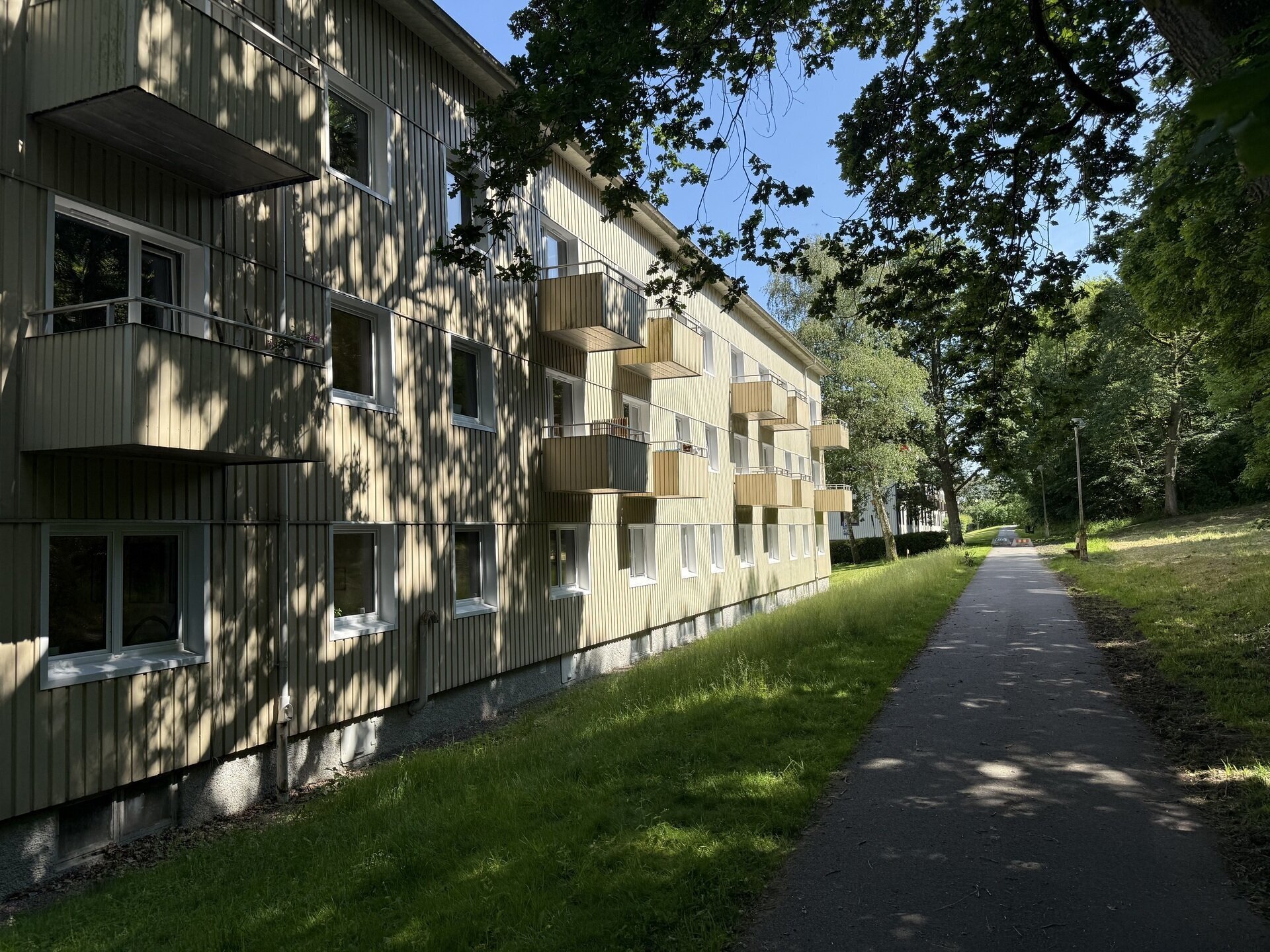 Lång rad flerbostadshus med balkonger och nya fönster längs gångväg i lövskog