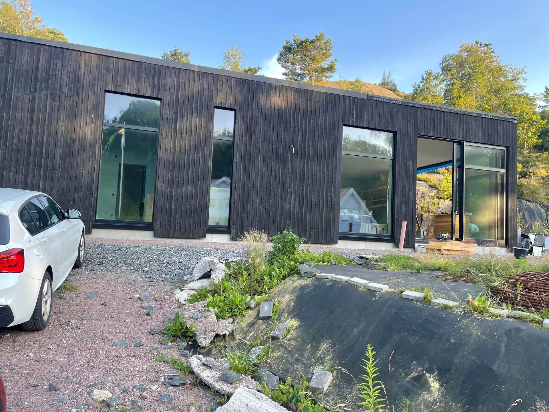 Mörk träpanelbyggnad med svarta fönsterpartier sedd från naturtomt med klippor