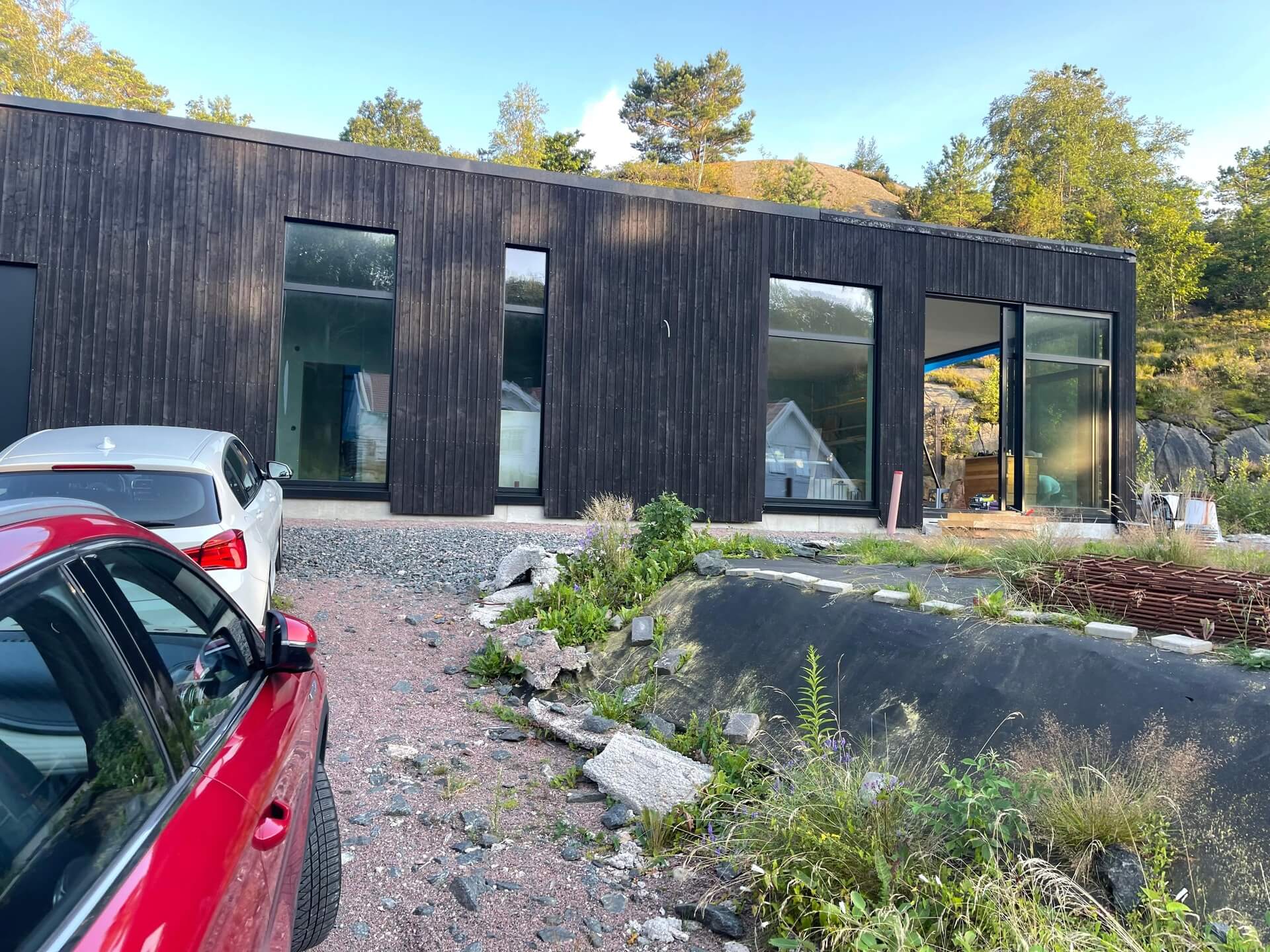Mörk träpanelbyggnad med stora svarta fönsterpartier mot naturtomt och klippa