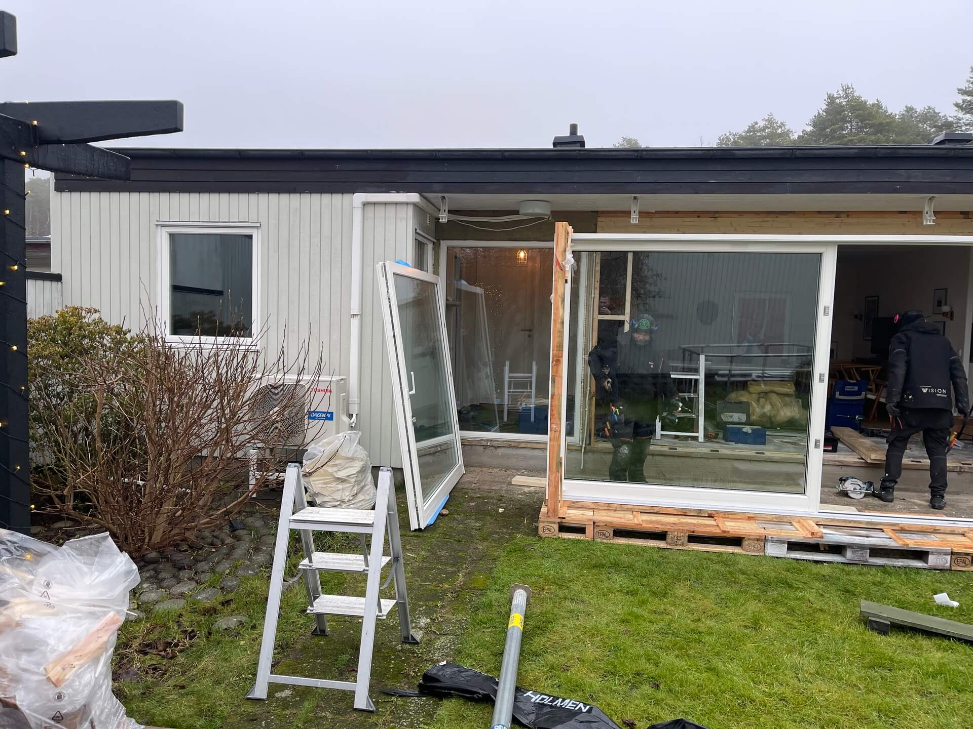 Gamla fönsterpartier uppställda i trädgård under pågående fönsterbyte på enplansvilla