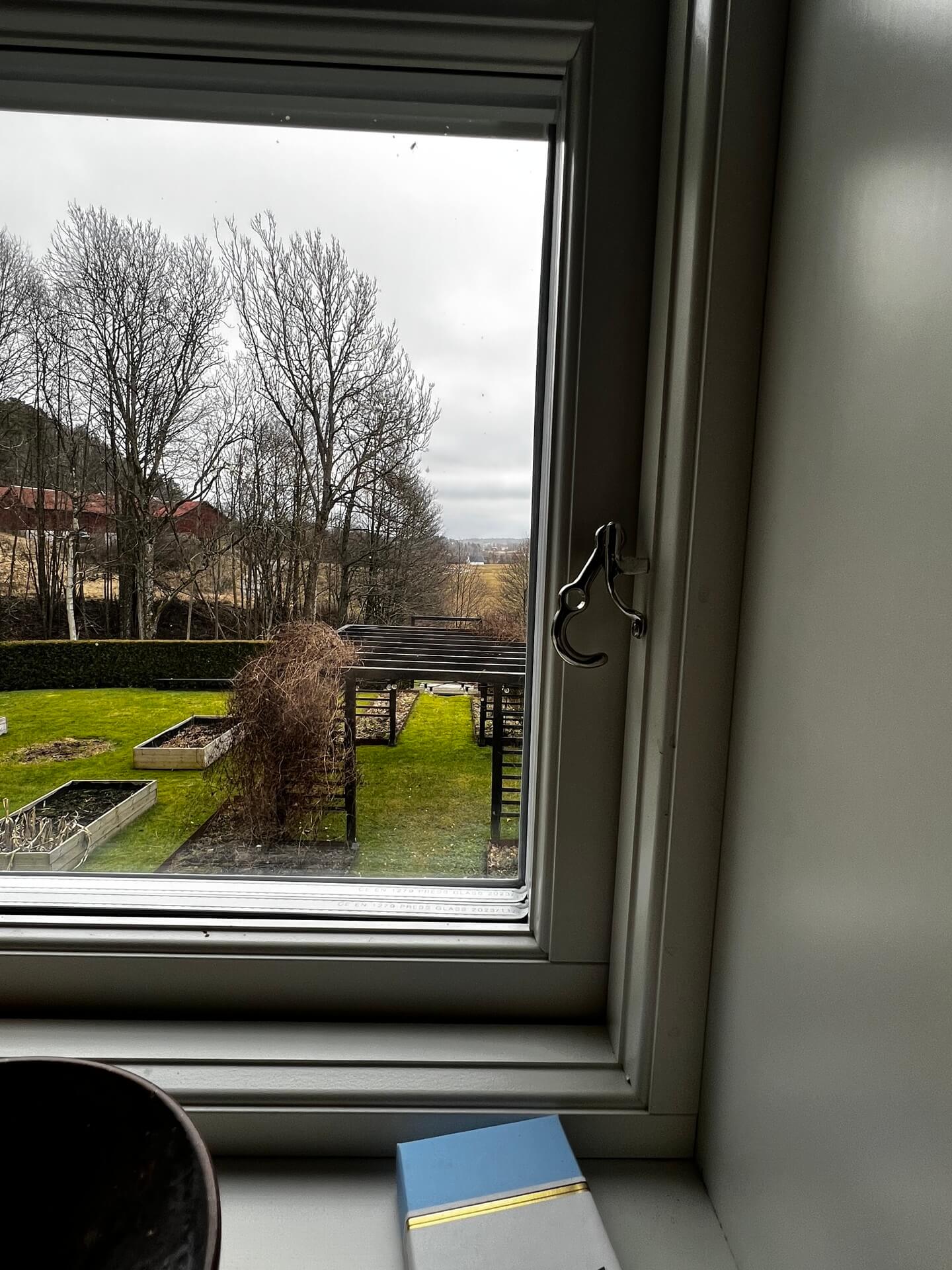 Öppnat sidohängt fönster med mörk karm och utsikt över trädgård med odlingslådor