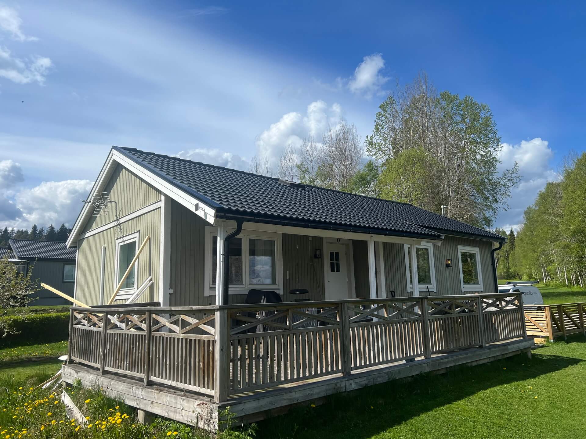Grågrön trävilla med svart pannat tak och stor veranda med träräcke en solig sommardag