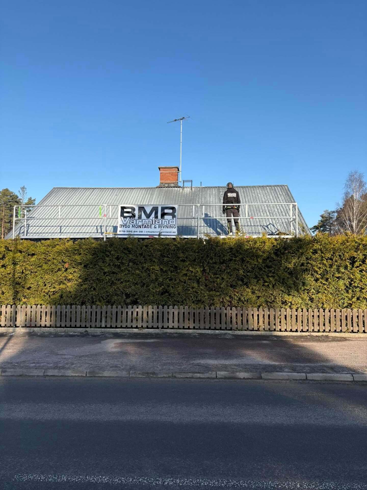 Hus med ställning längs takfoten och BMR Värmlands skylt, sett från gatan bakom häck
