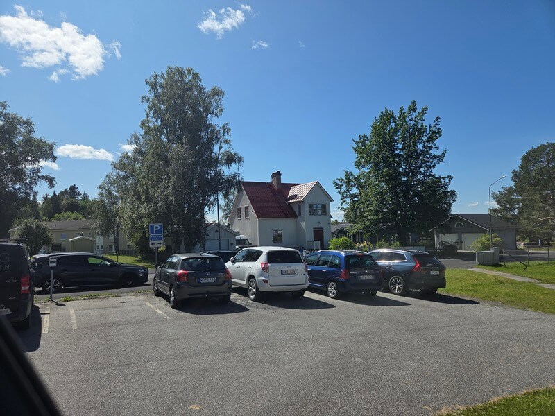 Vit villa med rött tak sedd från parkeringsplats i sommarmiljö
