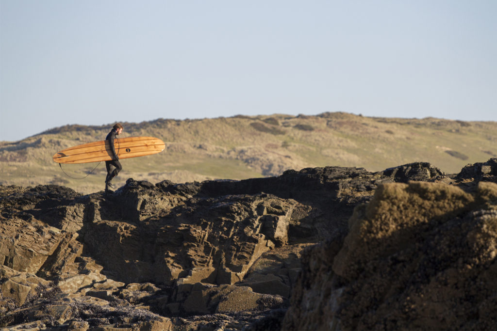 Chris walks back up the cliff with the Otter Surfboards wooden longboard Seasaw at Godrevy