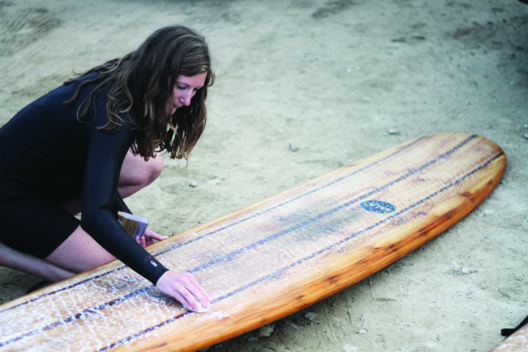 Rebecca Pepperell waxing the Otter Surfboards Seasaw wooden longboard at Perranporth in Cornwall