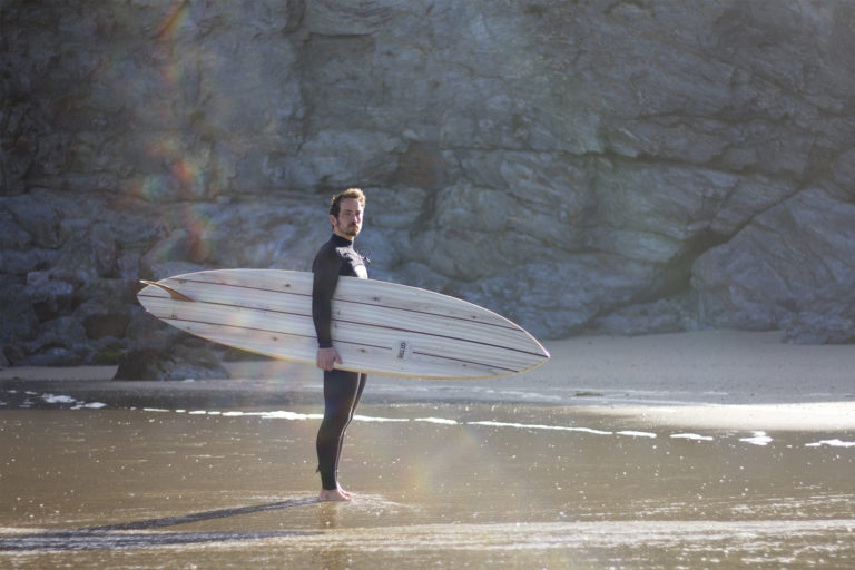 James Otter portrait, standing on the beach at porthtowan with his Clipper wooden surfboard.