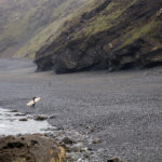 Johnny Leon walking out of the surf under the cliffs at Milook with his Clipper Otter wooden surfboard.