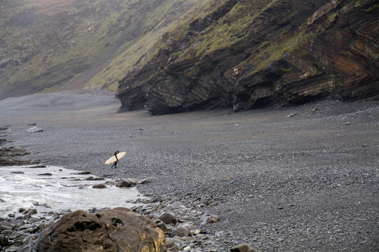 Johnny Leon walking out of the surf under the cliffs at Milook with his Clipper Otter wooden surfboard.