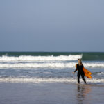 Sarah walks into the surf with the otter surfboards wood burner Wooden surfboard at Gwithian, Cornwall shot from the beach