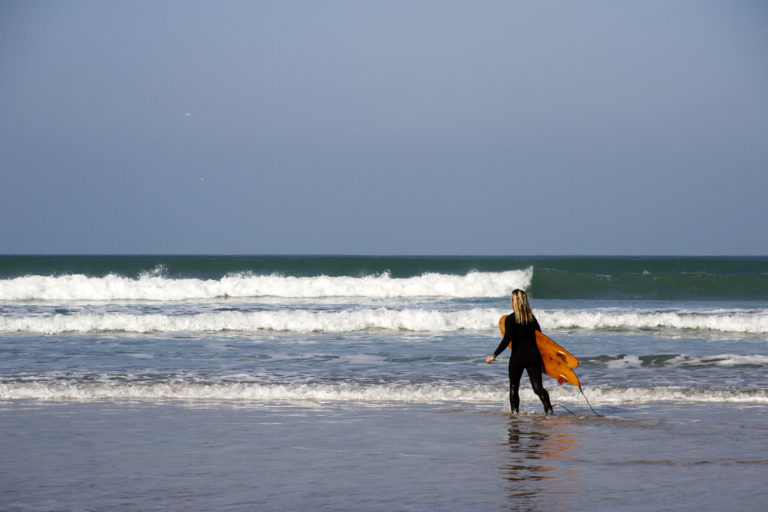 Sarah walks into the surf with the otter surfboards wood burner Wooden surfboard at Gwithian, Cornwall shot from the beach
