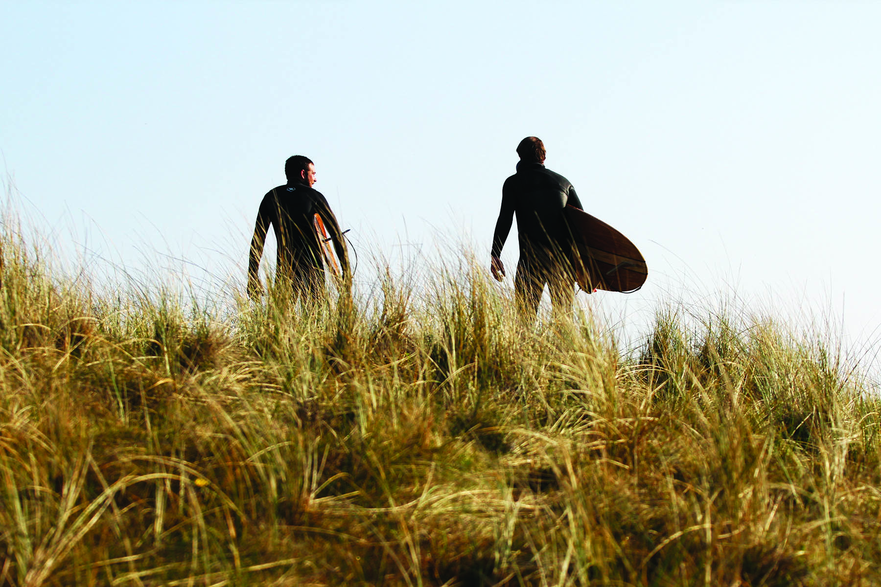 Chris and Gideon walk along the sand dunes in maram grass with Otter Surfboards wooden Island Hopper and Jetty at Gwithian