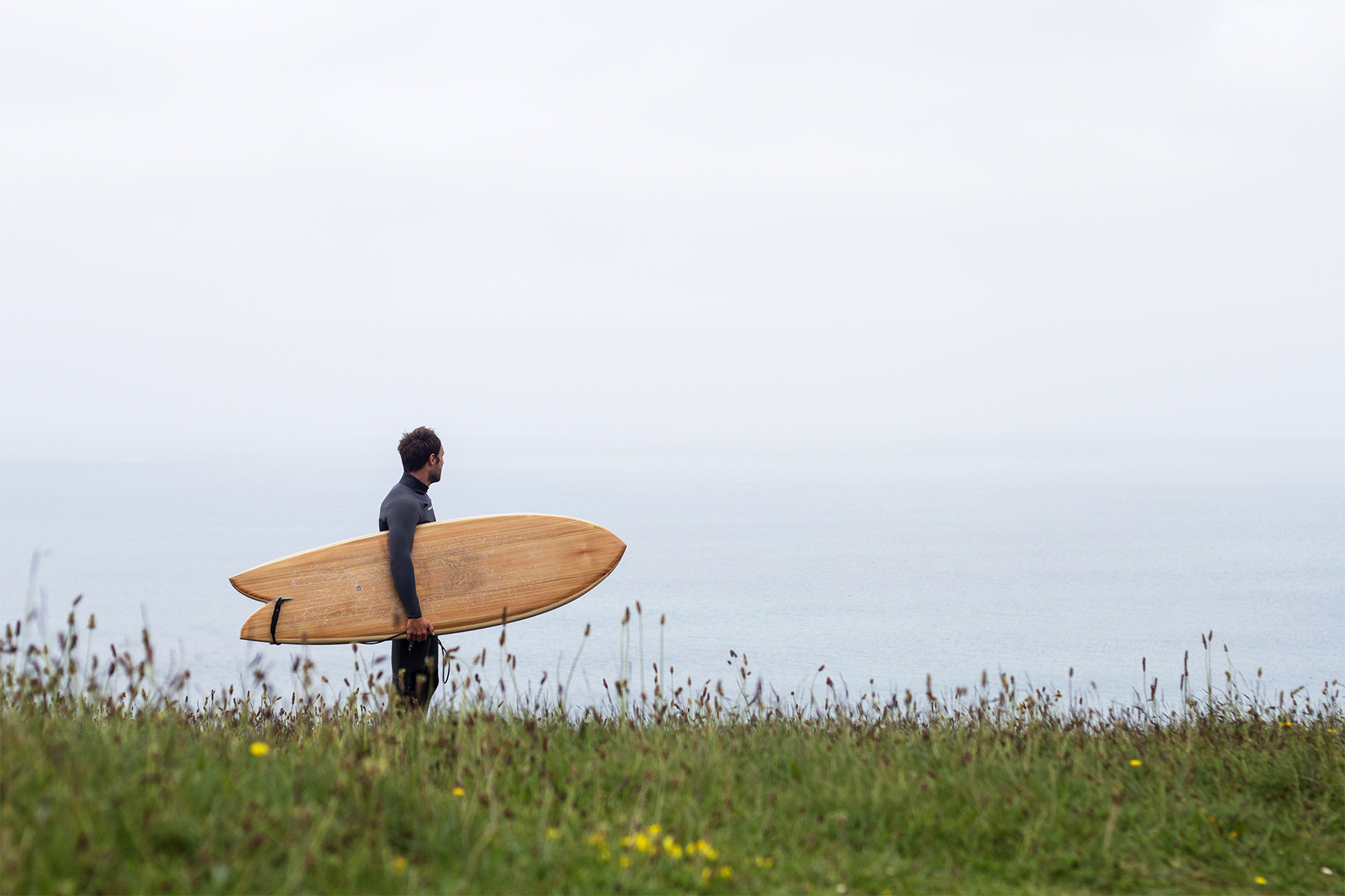 Ally Stands on the cliffs in the grassy meadow at Porthtowan with his Otter Wooden surfboard Woodburner.