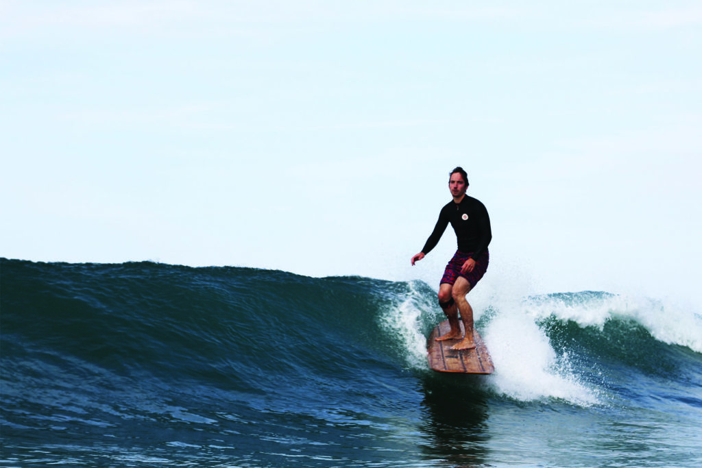 James Otter with the Pier wooden surfboard at Chape Porth, Cornwall in summer. Riz Boardshorts