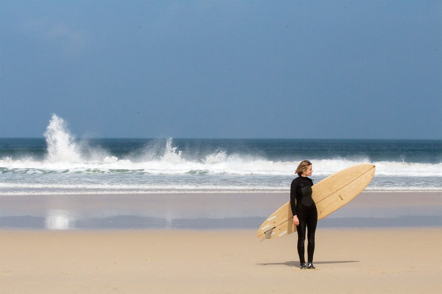 Sophie Hellyer on the beach in wetsuit with otter wooden surfboard coaster