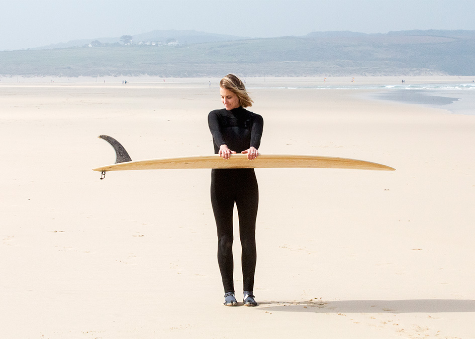 Sophie Hellyer on the beach in wetsuit with otter wooden surfboard coaster