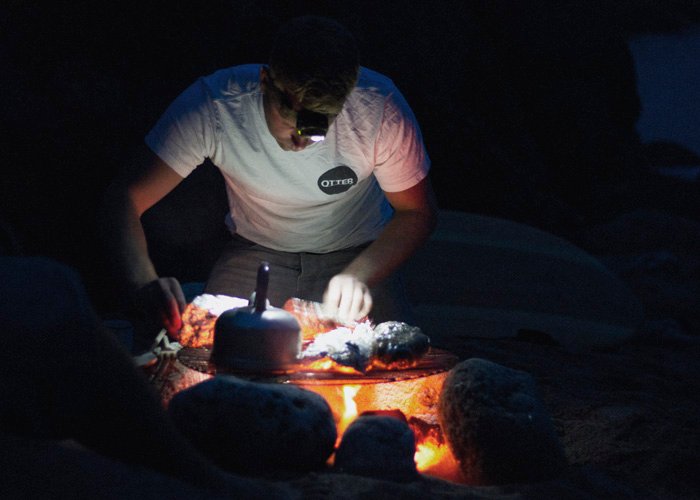 sean cooking on fire with wooden surfboard on the beach