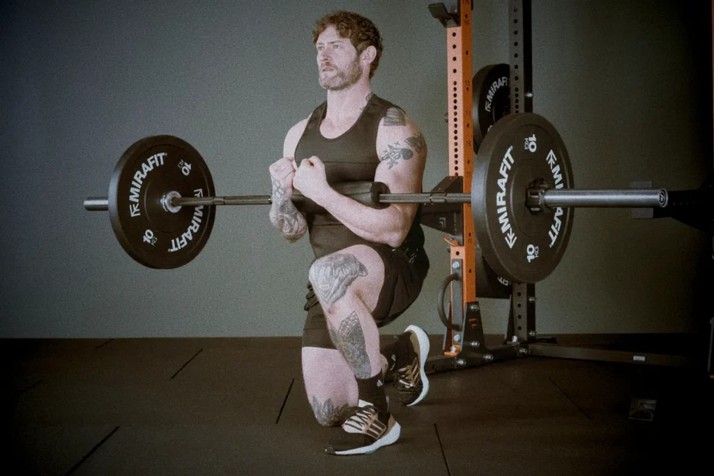 Athlete performing barbell Zercher lunge, bar held in elbows close to chest, front leg bent, torso upright, cinematic gym lighting showing balance and stability