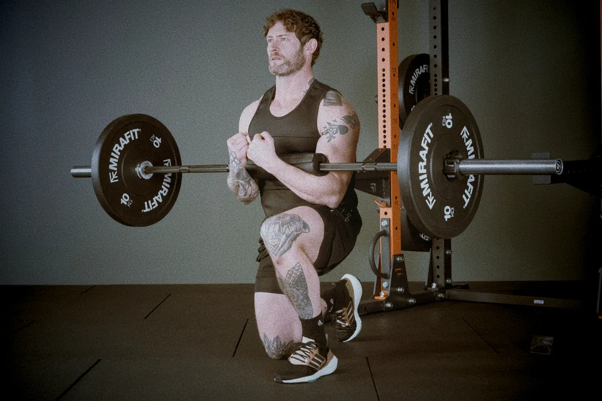 Athlete performing barbell Zercher lunge, bar held in elbows close to chest, front leg bent, torso upright, cinematic gym lighting showing balance and stability