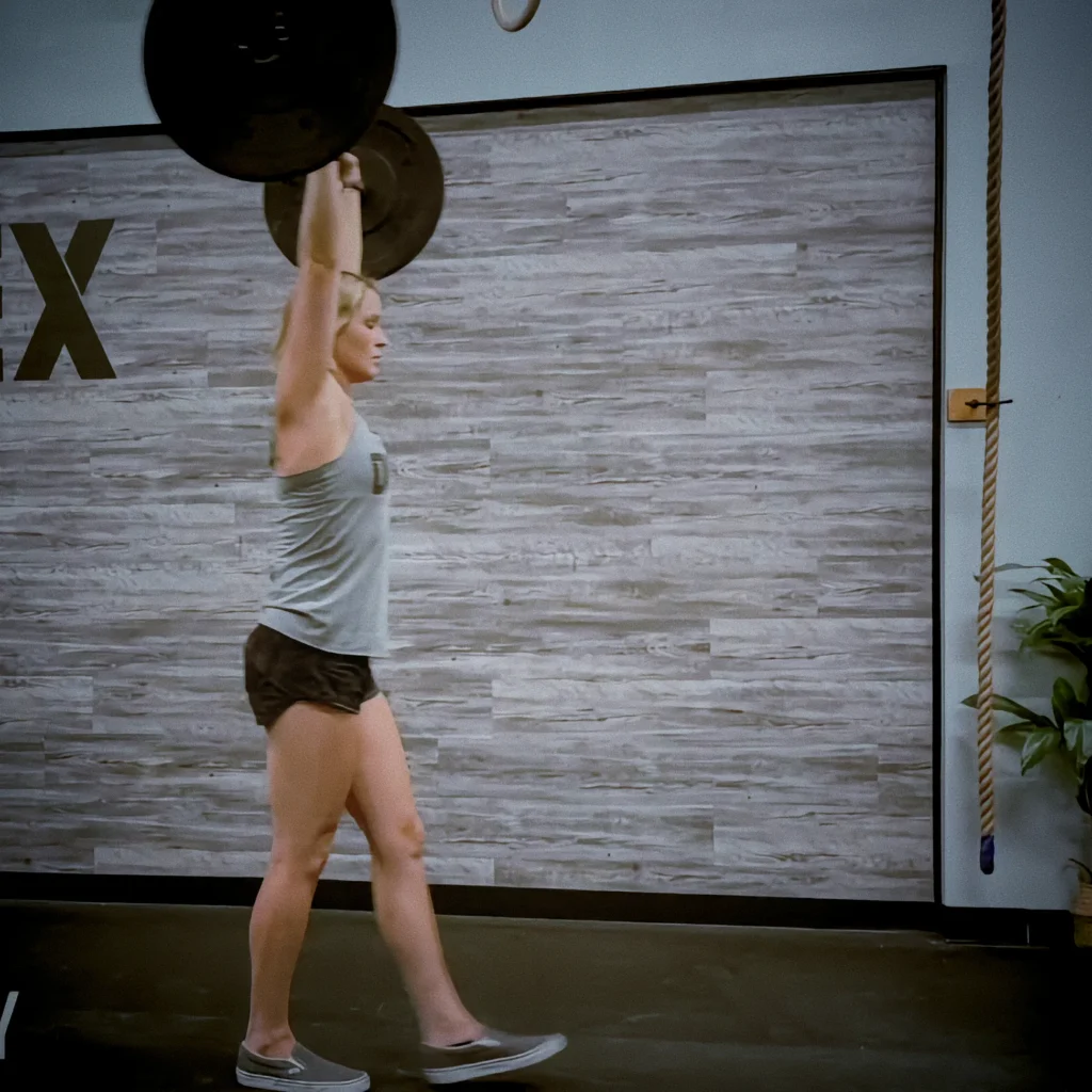 Athlete performing barbell overhead carry, bar locked overhead, upright torso, focused expression, cinematic gym lighting showing control, balance, and strength