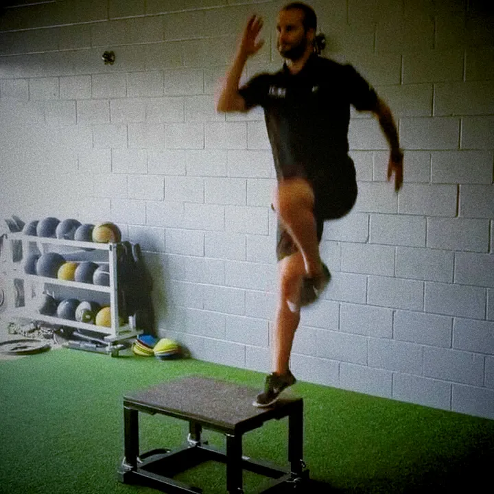 Athlete performing explosive step-up on plyo box, mid-air with front leg extended and rear knee driving upward, cinematic gym lighting showing power and balance