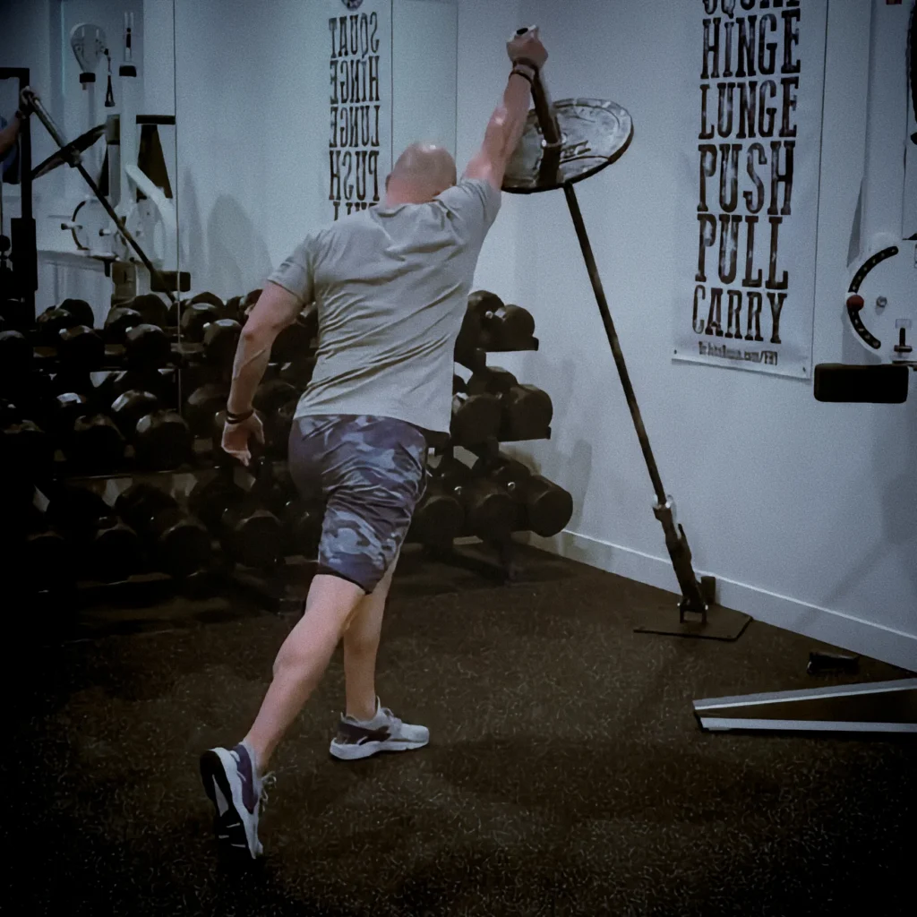 Athlete performing landmine rotational press, mid-pivot with hips turned and barbell pressing diagonally overhead, cinematic gym lighting