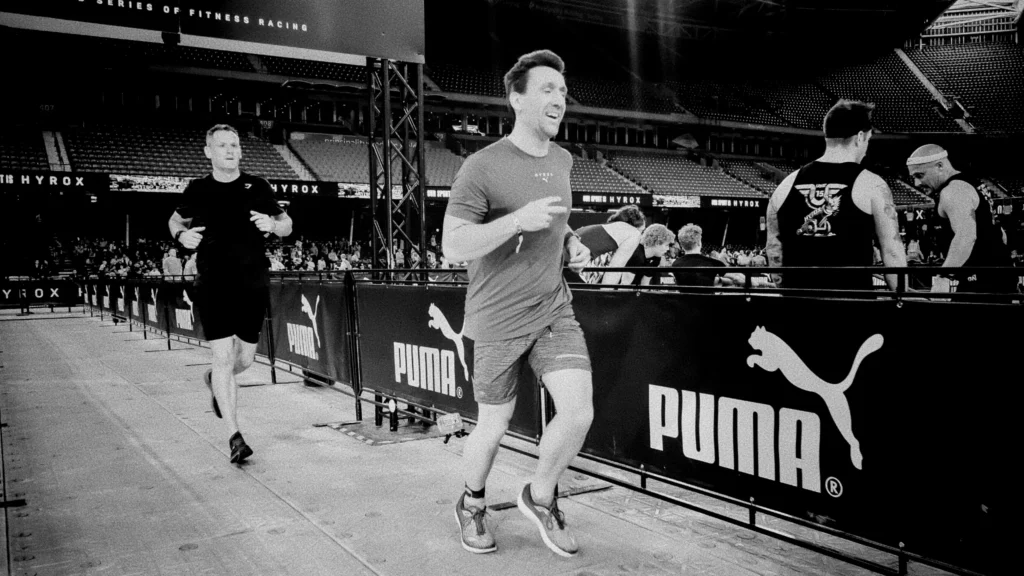 Two HYROX partners tagging between stations in Doubles race, smiling but fatigued