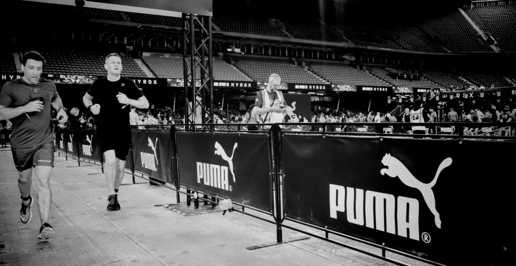 HYROX doubles team mid-run side by side, synchronised stride, cinematic warm lighting and background crowd