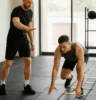 Coach guiding an athlete through a balance drill or movement pattern (e.g. crawling, single-leg balance, agility ladder) in a light-filled gym or outdoor setting