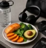 Plate of vibrant, whole foods next to gym gear e.g. water bottle, lifting straps, headphones on a kitchen counter or gym bag