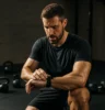 Athlete checking a timer or stopwatch mid-workout, surrounded by kettlebells, dumbbells, or functional fitness equipment