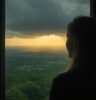 A contemplative woman stands at a window, watching the contrast of storm and sunlight, reflecting the changeable nature of emotions